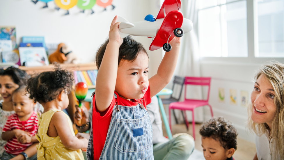 (Quelle: Rawpixel.com - stock.adobe.com) Kleinkind spielt mit Spielflugzeug aus Holz inmitten einer Kindergartengruppe