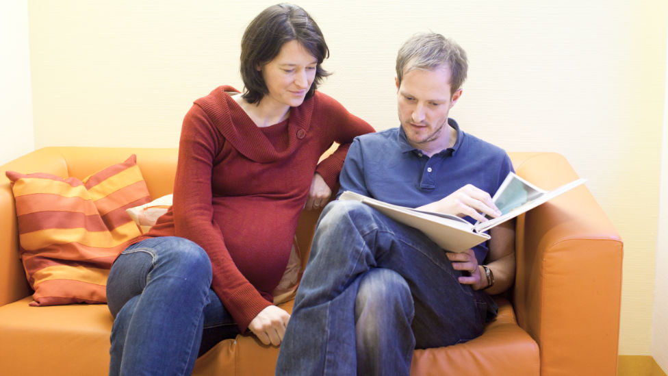 Schwangeres Paar sitzt gemeinsam auf einem Sofa im Wartebereich und liest eine Zeitschrift