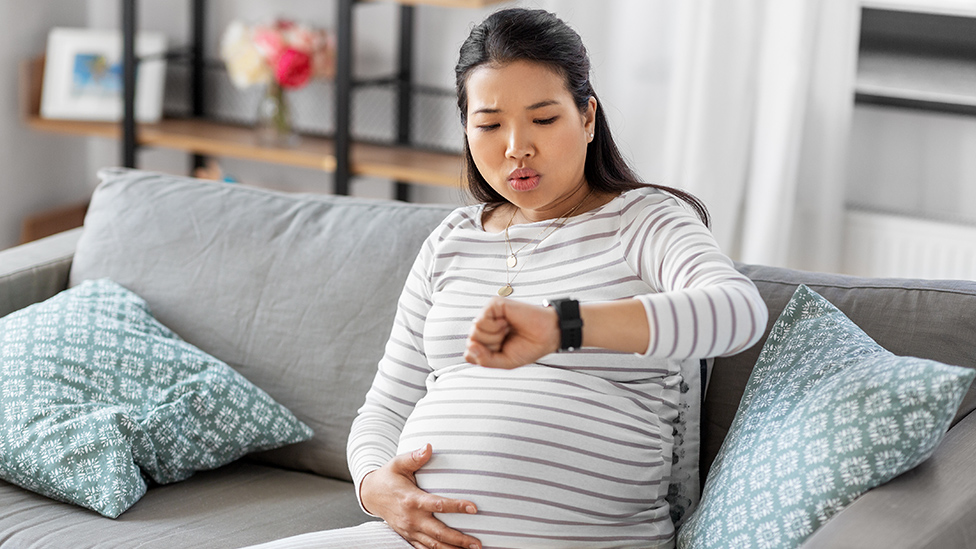 Schwangere Frau hält eine Hand an den Bauch, schaut auf ihre Armbanduhr an der andere Hand und atmet bewusst aus