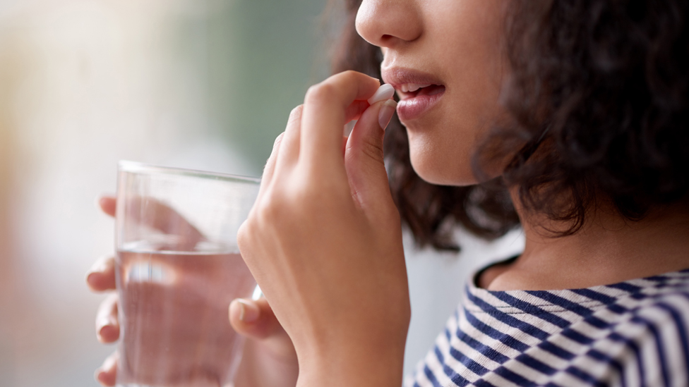 Die Pille durchgehend einnehmen (Quelle: iStock.com/Tassil) Frau mit schwarzen lockigen Haaren nimmt weiße Pille ein mit Wasserglas in der rechten Hand